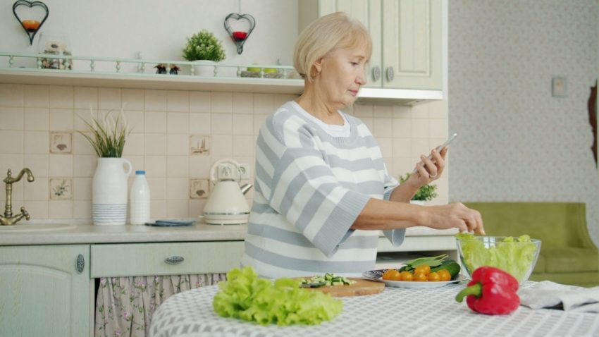 Old lady is cooking vegetable salad and looking at smartphone screen checking ingredients for meal during dinner preparation at home. People and gadgets concept.